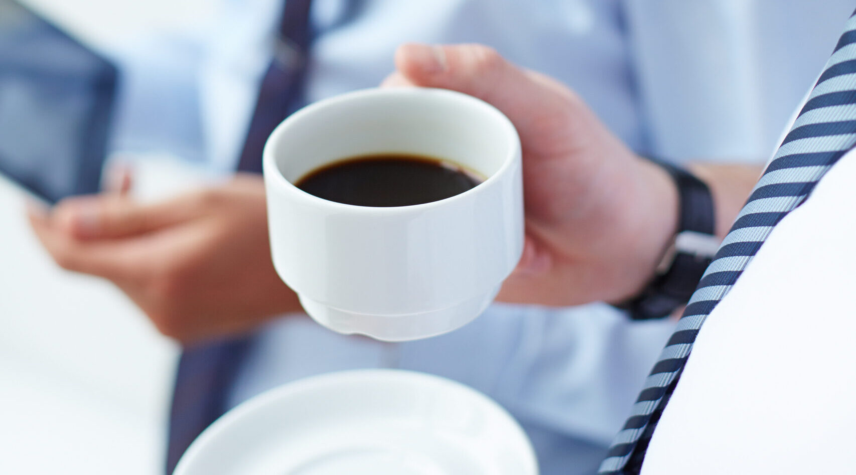 Image of young businessman holding cup of coffee while listening to his colleague at meeting